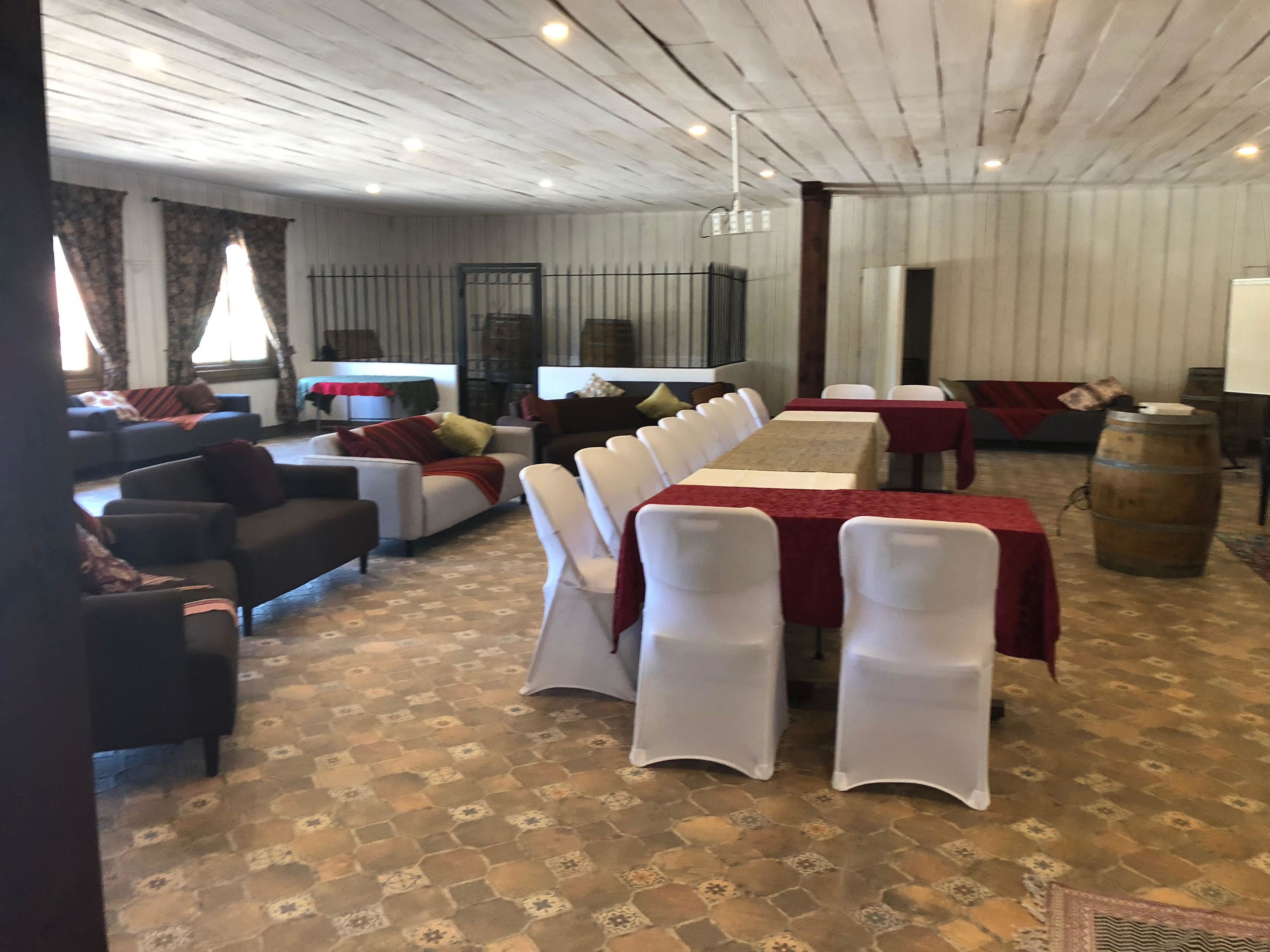 Uma sala de jantar com uma mesa comprida coberta por um tecido vermelho, rodeada por cadeiras brancas. Há sofás e uma pequena mesa com um tecido vermelho ao fundo. O quarto possui um chão estampado e teto de madeira.