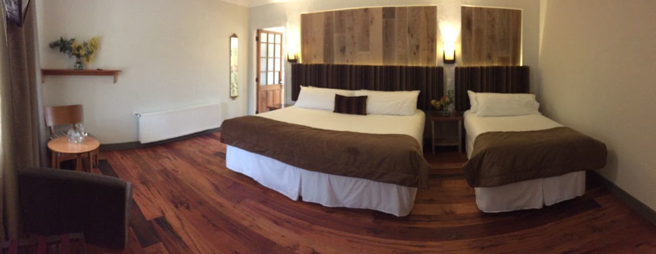 Habitación de hotel con dos camas, piso de madera, una mesita con una silla y un estante en la pared con flores.
