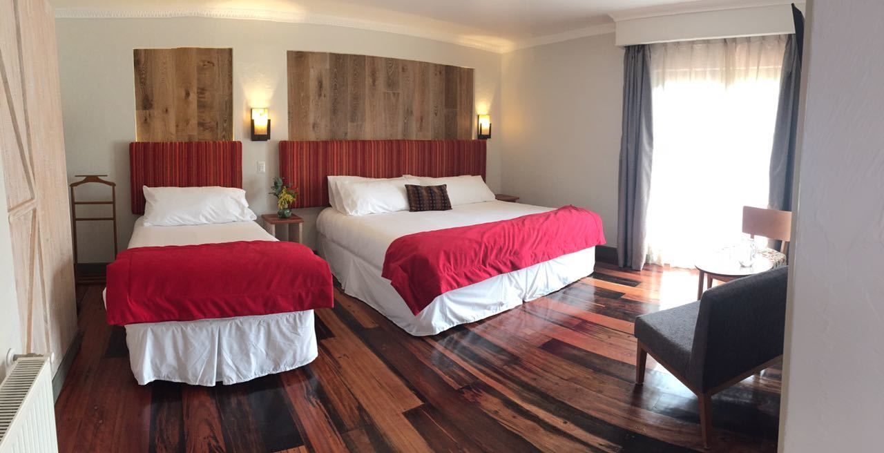 Quarto de hotel com duas camas, cabeceira de madeira, uma mesinha e uma cadeira.