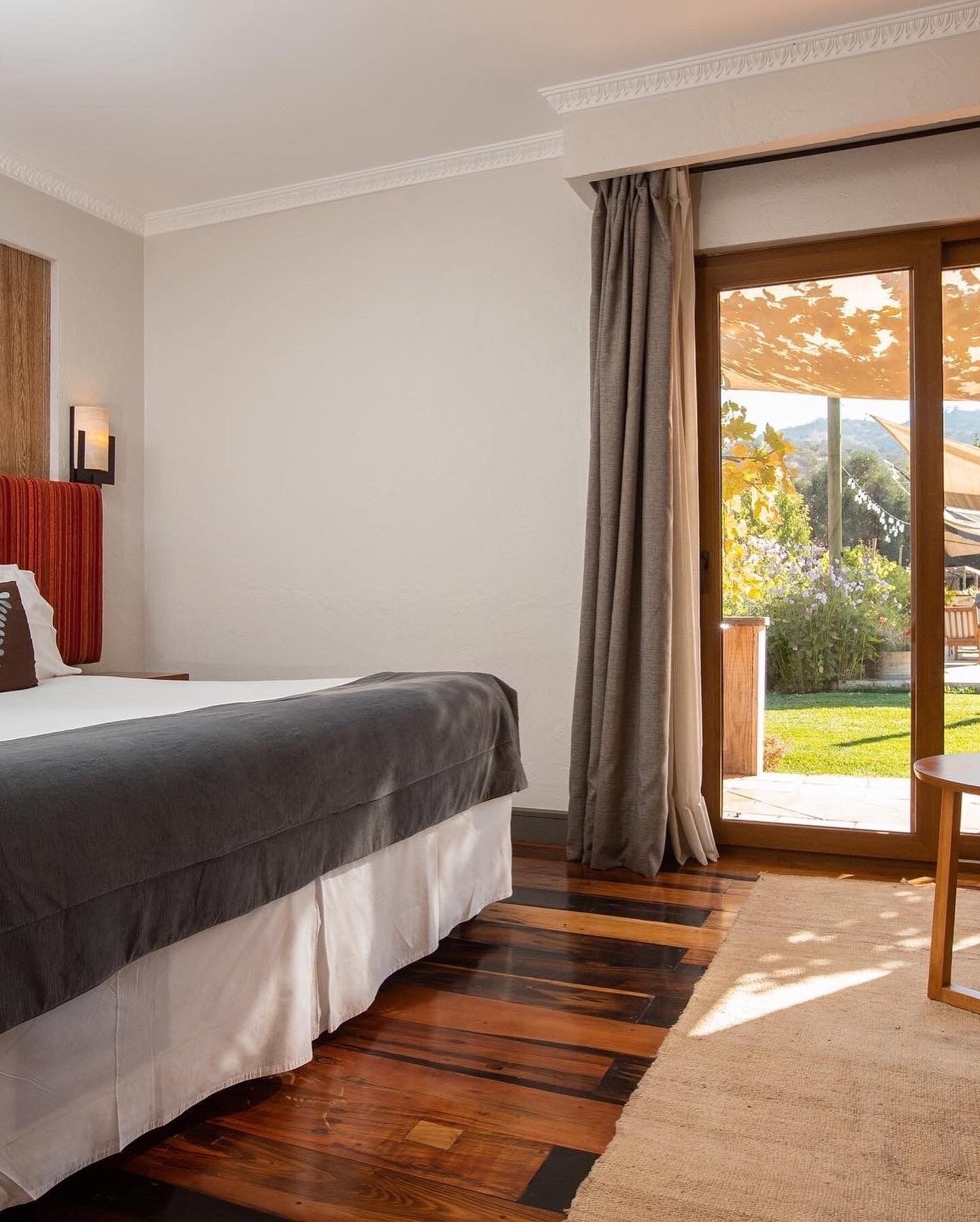 Un dormitorio con una cama grande, piso de madera, vista a un jardín por una puerta de cristal y una mesita.