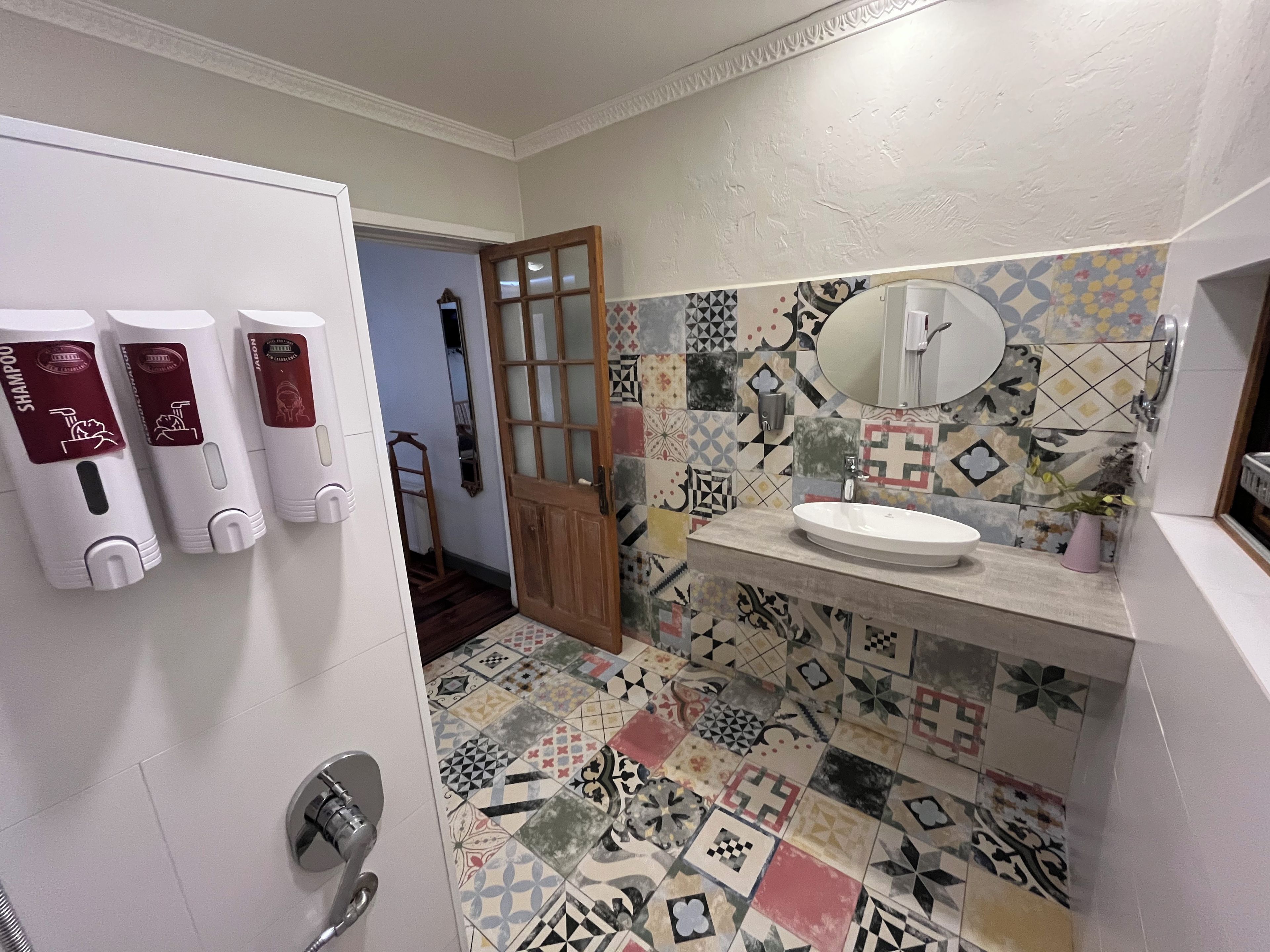 Un baño con suelo de baldosas, lavabo, espejo y tres dispensadores en la pared.