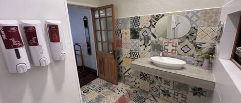 Un baño con suelo de baldosas, lavabo, espejo y tres dispensadores en la pared.