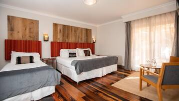 A hotel room with two beds, a wooden headboard, a small table with chairs, and a window with curtains.