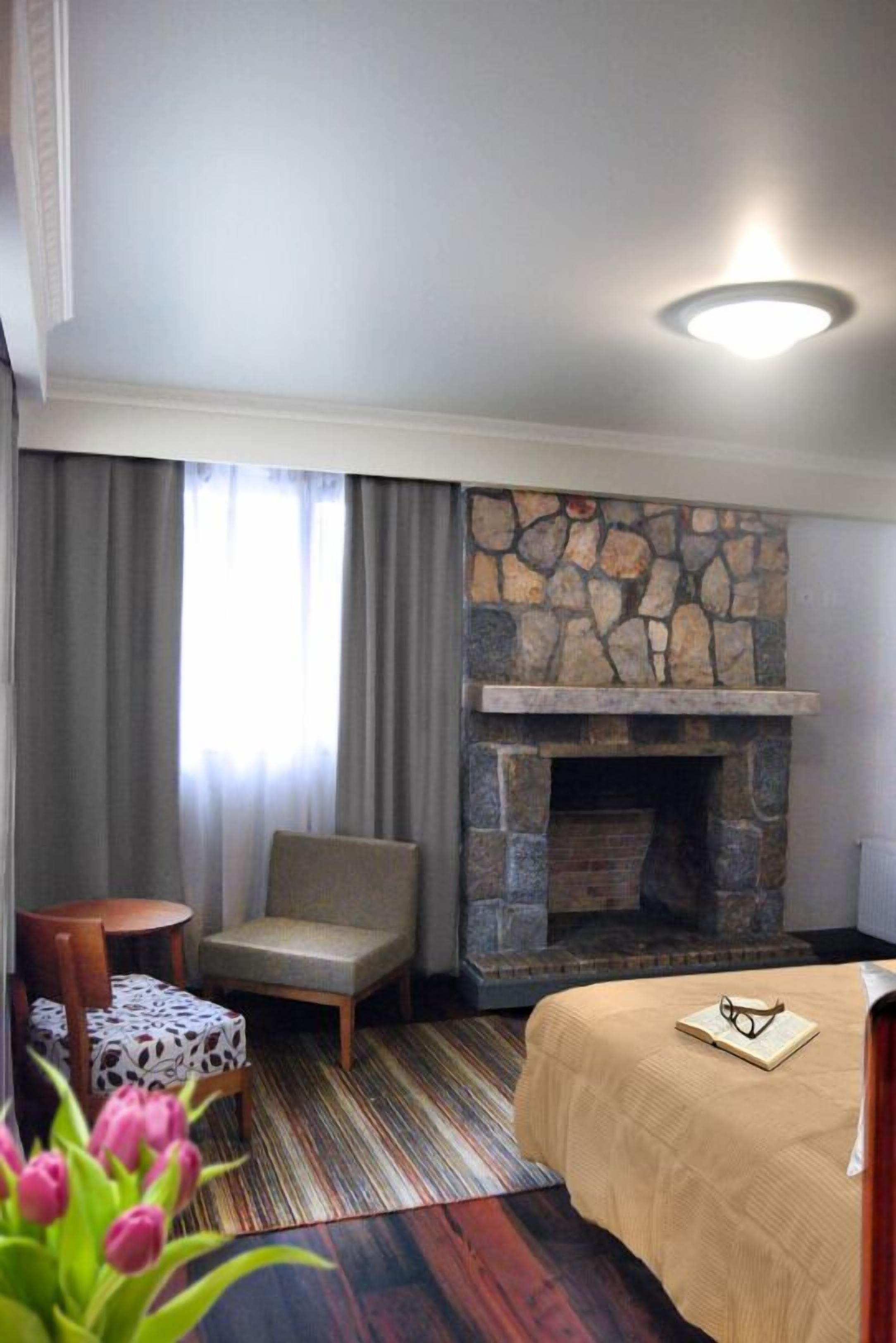 Une chambre d’hôtel avec une cheminée en pierre, un lit recouvert d’une couverture beige, une petite table avec une nappe à motifs, une chaise et une fenêtre avec des rideaux.
