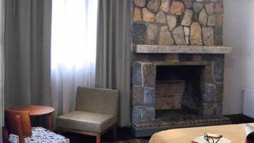 Une chambre d’hôtel avec une cheminée en pierre, un lit recouvert d’une couverture beige, une petite table avec une nappe à motifs, une chaise et une fenêtre avec des rideaux.