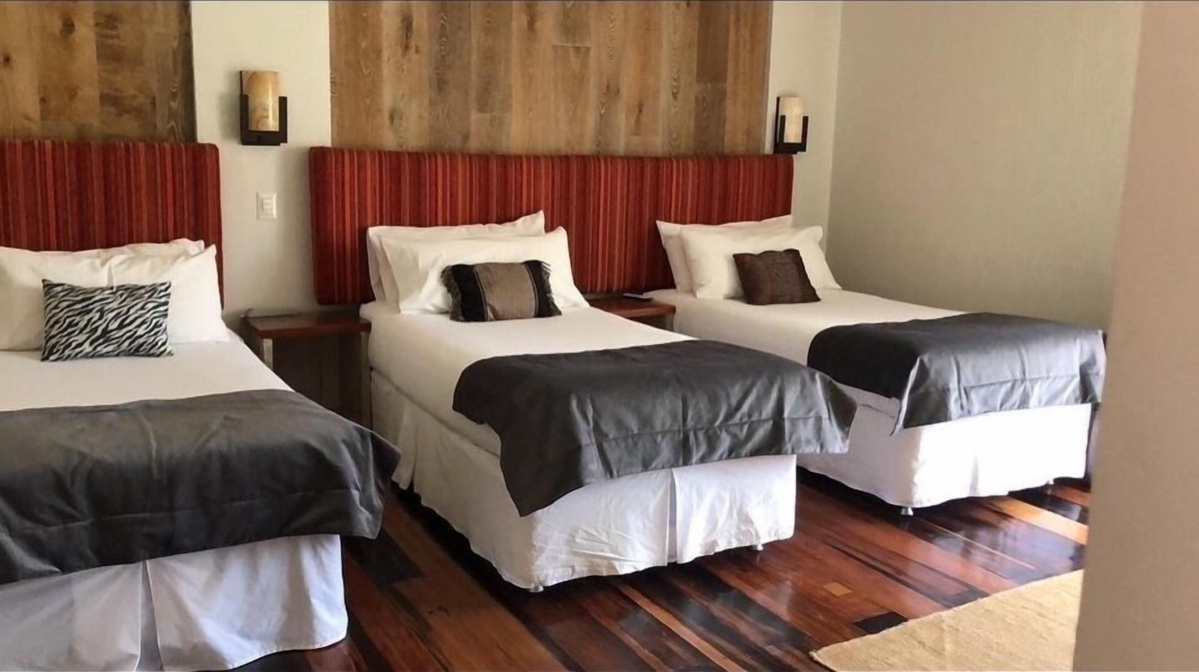 A hotel room with two single beds, each with a headboard, a nightstand, and a lamp. The room has wooden flooring and a large window.