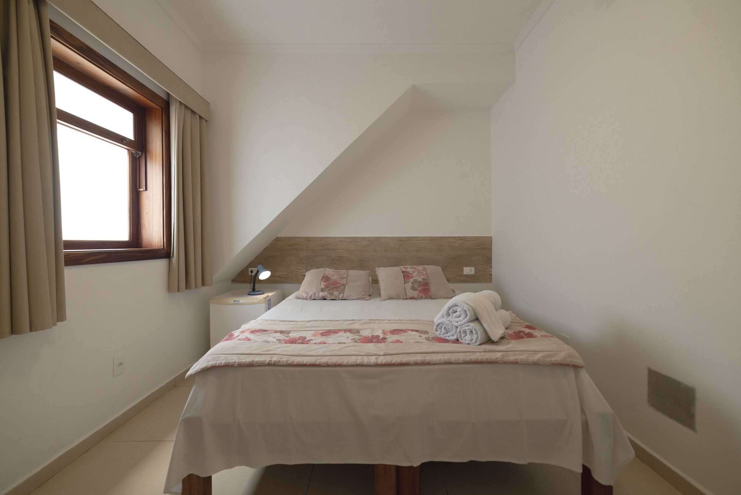 Quarto com cama, mesa de cabeceira, abajur e janela com cortinas.
