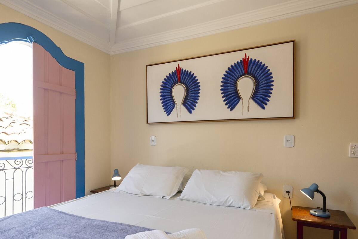 Un lit bien fait, recouvert d’une couverture bleue, avec des oreillers blancs et une serviette blanche pliée posée sur le dessus. Au-dessus du lit, on aperçoit une œuvre d’art encadrée, une petite table de chevet avec une lampe, et une porte dont le cadre est rose.