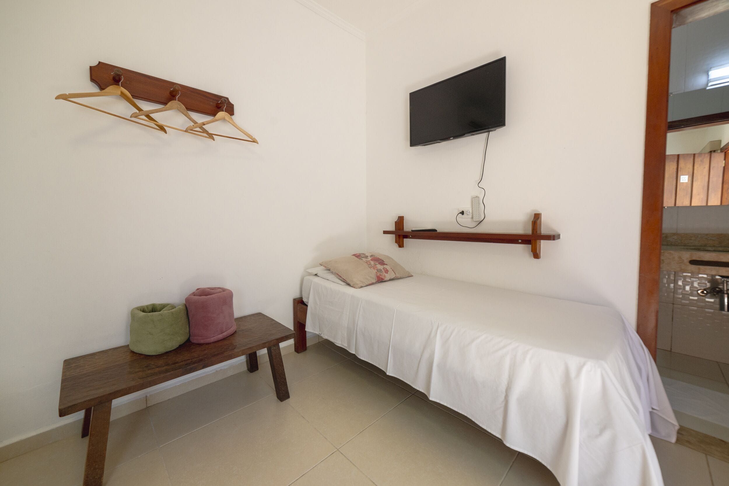 A small, simple room with a bed, a bench, a TV, and a wall-mounted TV stand.