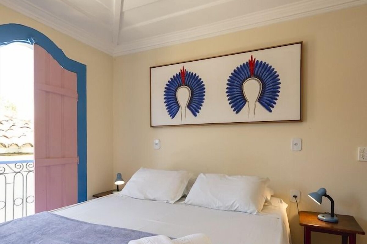 Un lit bien fait, avec une couverture grise, des oreillers blancs et une serviette blanche pliée posée sur le lit. On aperçoit une petite table de chevet avec une lampe, et au-dessus du lit, une œuvre d’art encadrée.