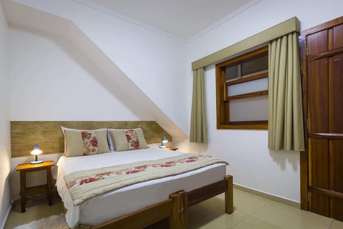 Une chambre avec un plafond en pente, un lit recouvert d’une couvre-lit à motifs floraux, deux tables de chevet avec des lampes, une fenêtre avec des rideaux et une armoire en bois.
