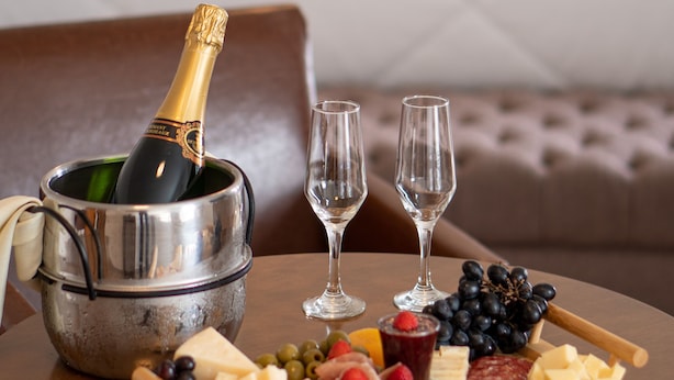 A bedroom with a bed, a bedside table with a bottle of champagne, two glasses, and a cheese board with various cheeses and fruits. - Pousada Itália Moderna (Campos do Jordão)