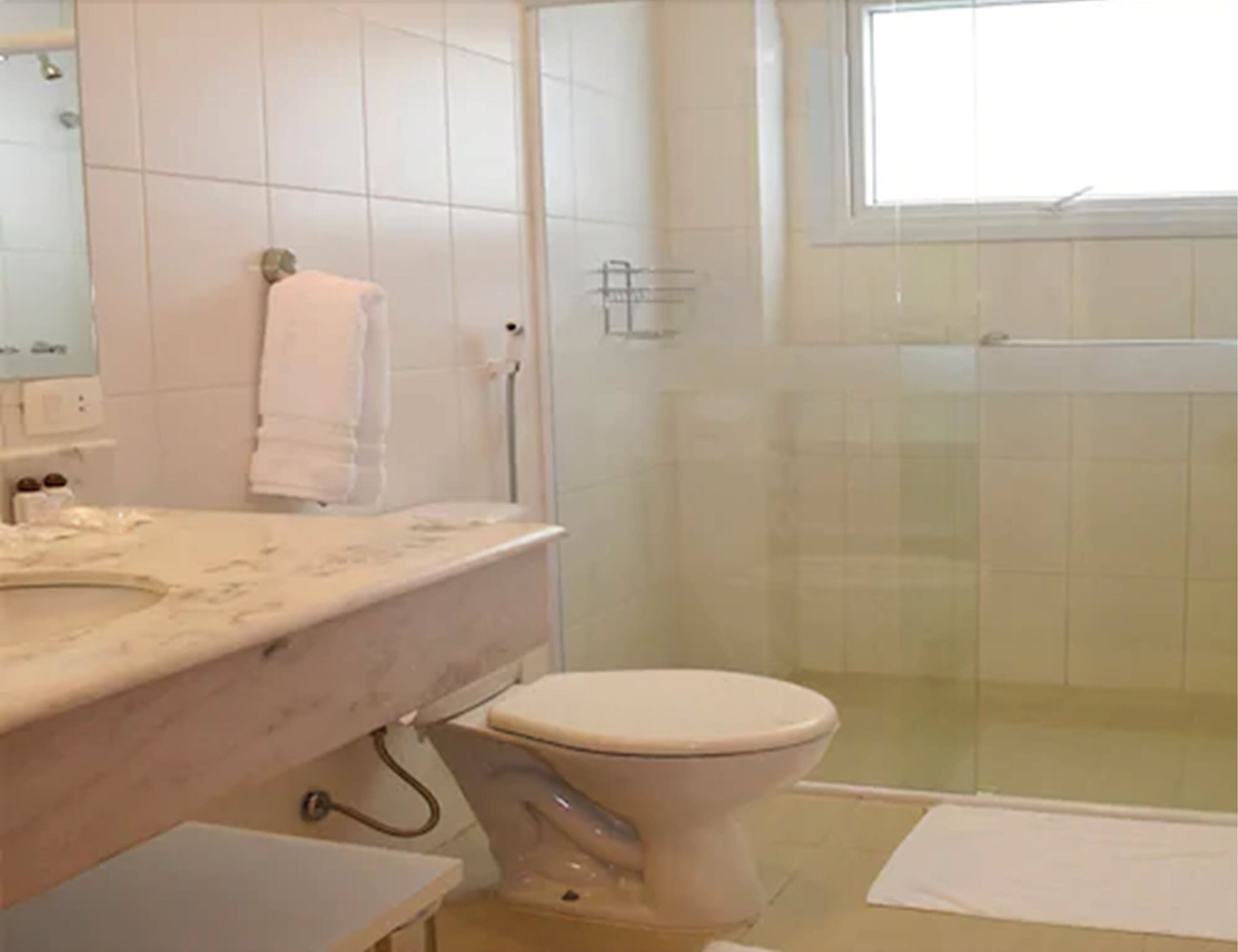 A bathroom with a white sink, mirror, and toilet. There is a glass shower enclosure and a window.