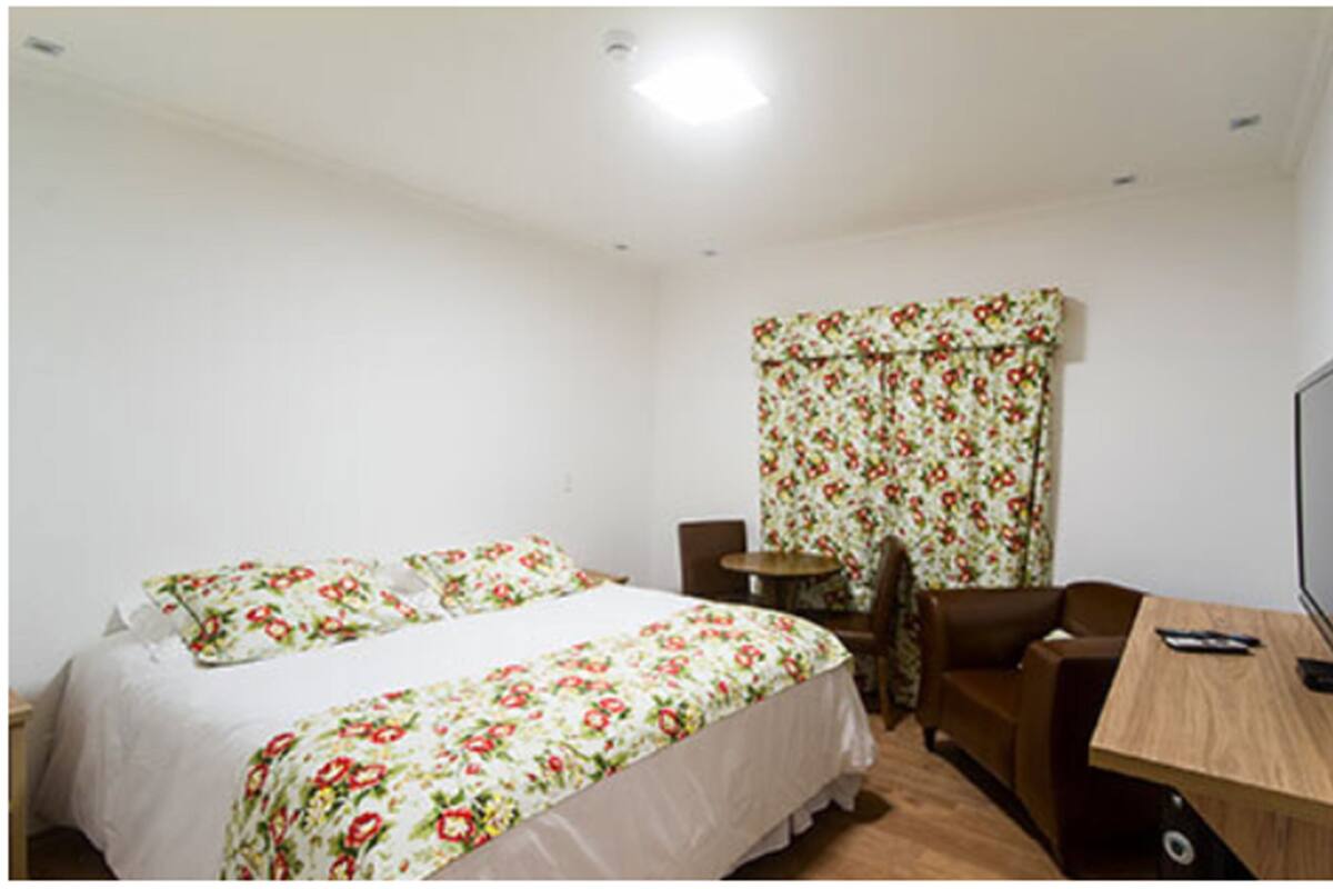 A hotel room with two beds, a desk, a chair, a TV, and a window with floral curtains.