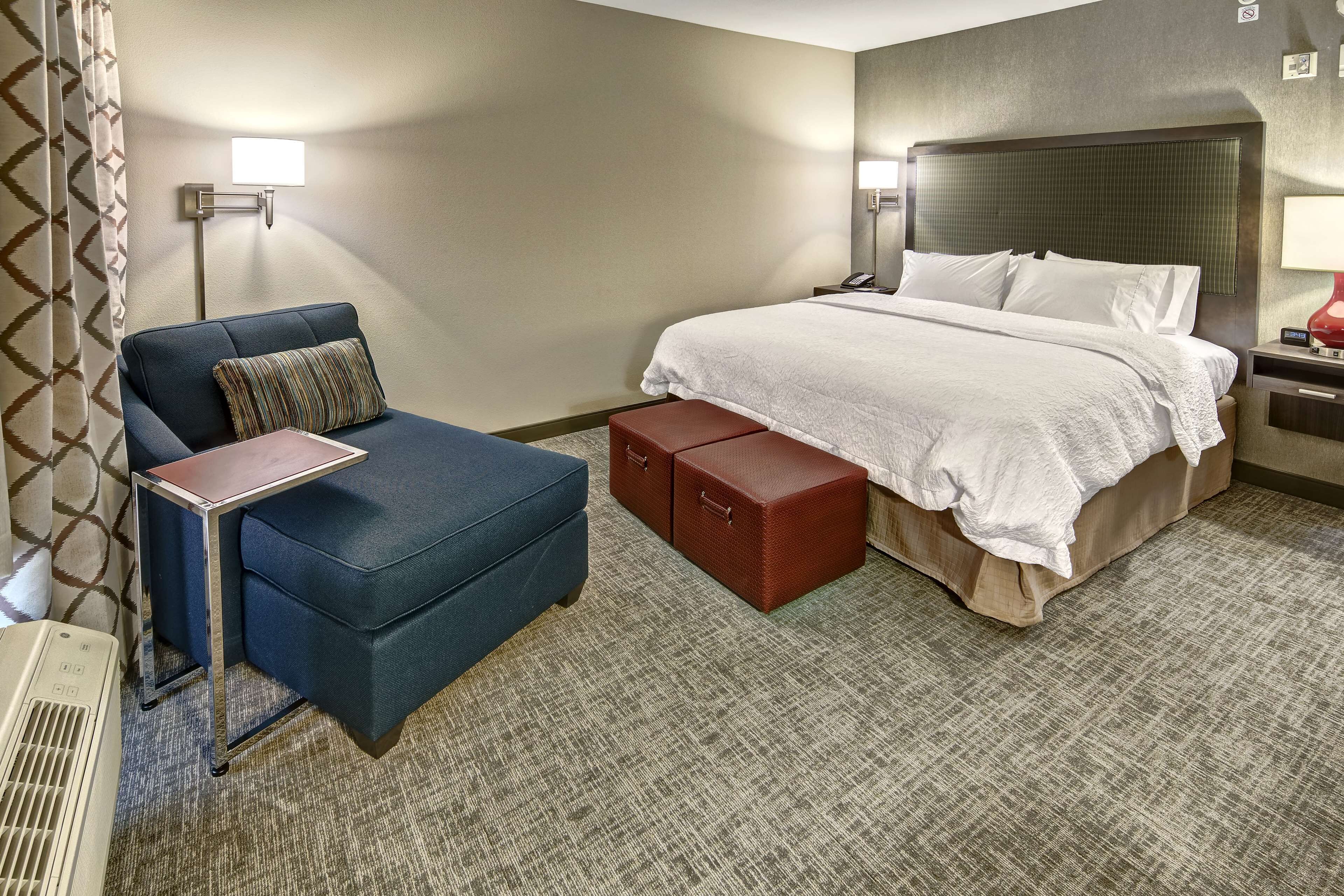 A hotel room with a bed, a blue armchair, a small table, and a red storage box.