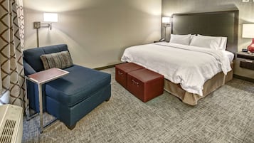 A hotel room with a bed, a blue armchair, a small table, and a red storage box.