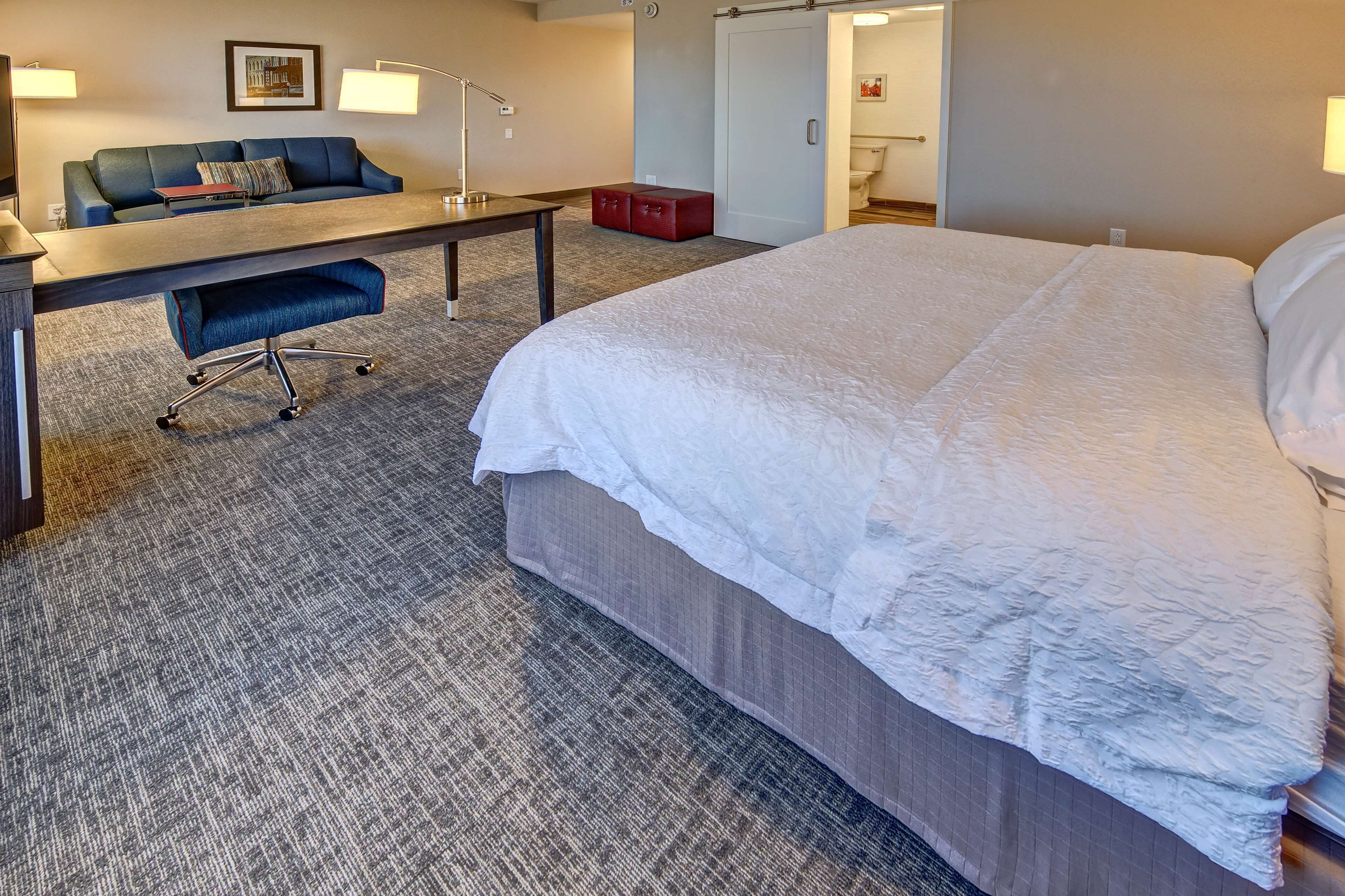 Una habitación de hotel con una cama grande, un escritorio con silla, un sofá y un pequeño mueble de almacenamiento rojo.