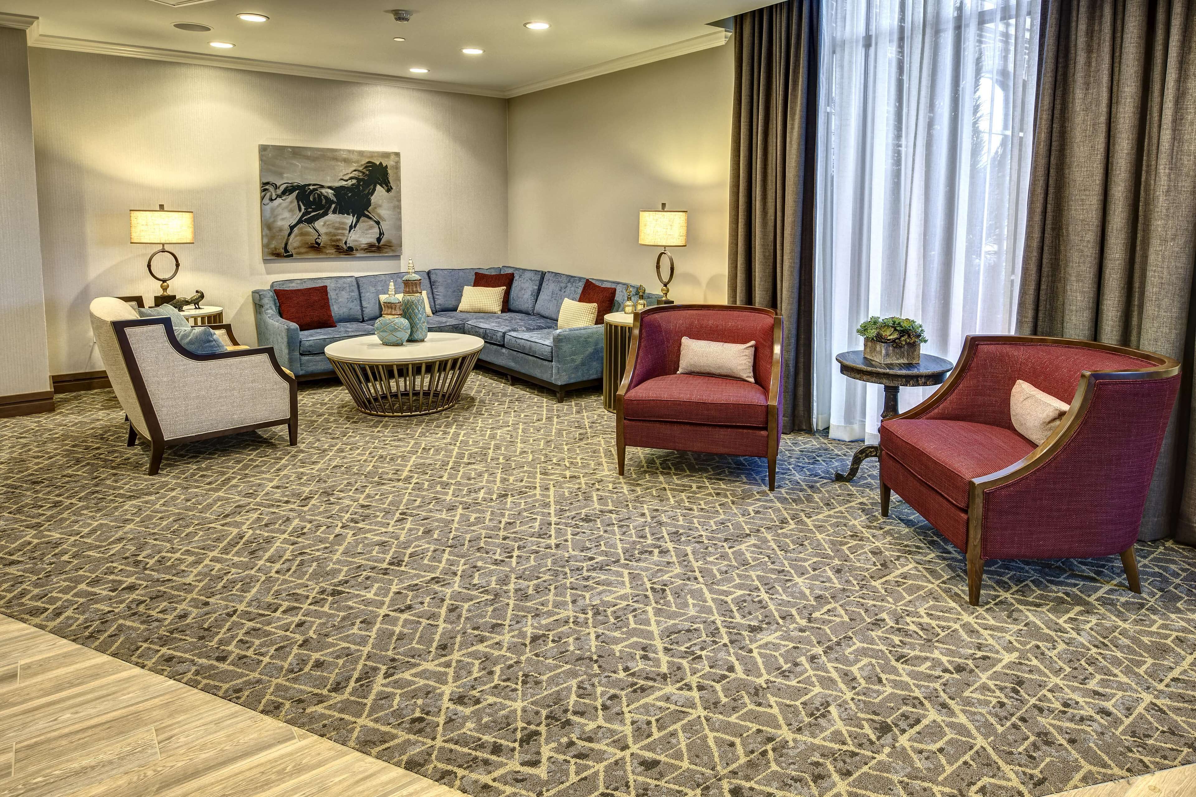 A hotel room with a patterned carpet, a blue sofa set, a red armchair, a small round table with a lamp, and a large window with curtains.