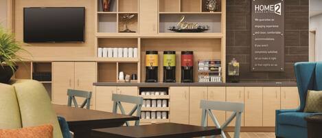 A modern hotel room with a flat-screen TV mounted on the wall, a coffee station with decaf and gourmet options, and a sign stating a home guarantee.