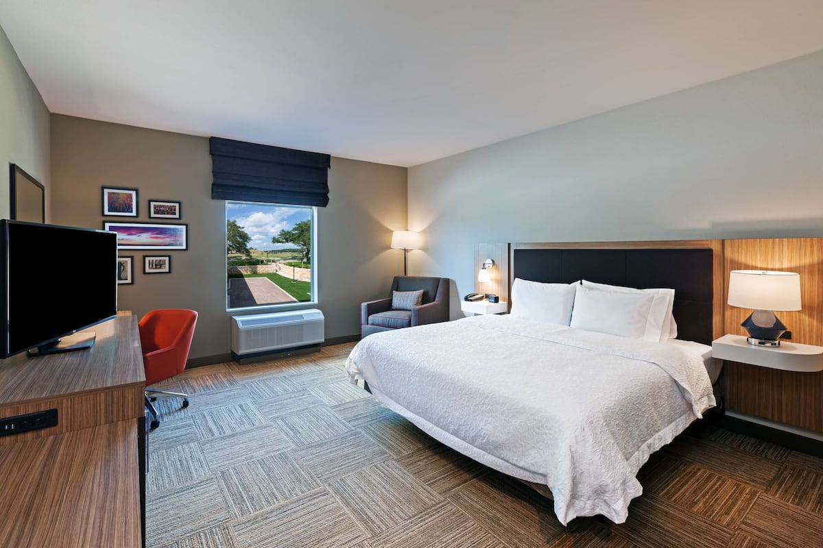 A hotel room with a large bed, a desk with a TV, a chair, a lamp, and a window with a view of the outdoors.