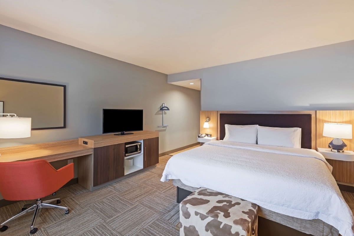 A hotel room with a bed, a desk with a TV, a red chair, and a nightstand with a lamp.
