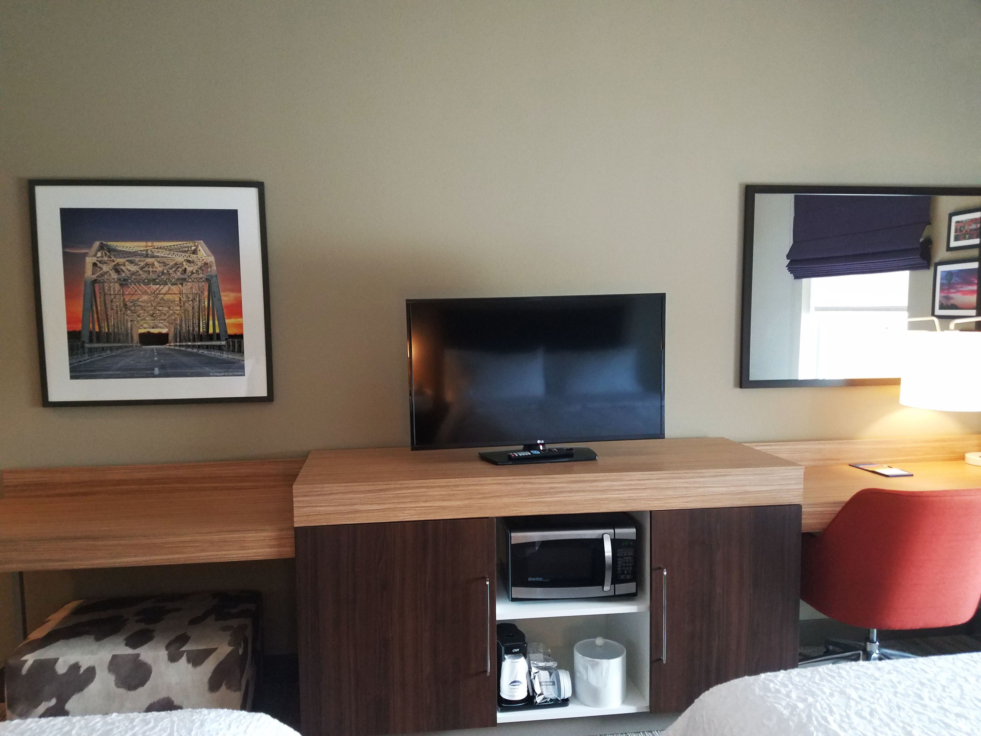 Una camera d'albergo con un letto, una TV su un mobile in legno, un forno a microonde, una sedia rossa e un quadro con il soggetto di un ponte.