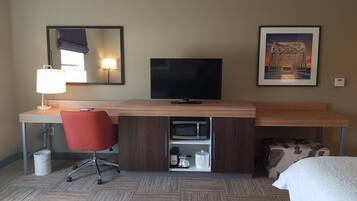 A hotel room with a wooden desk, a red office chair, a flat-screen TV, a microwave, a coffee maker, and a framed picture of a bridge.