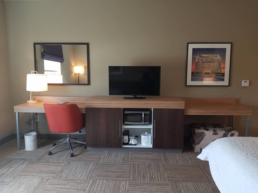 A hotel room with a wooden desk, a red office chair, a flat-screen TV, a microwave, a coffee maker, and a framed picture of a bridge.