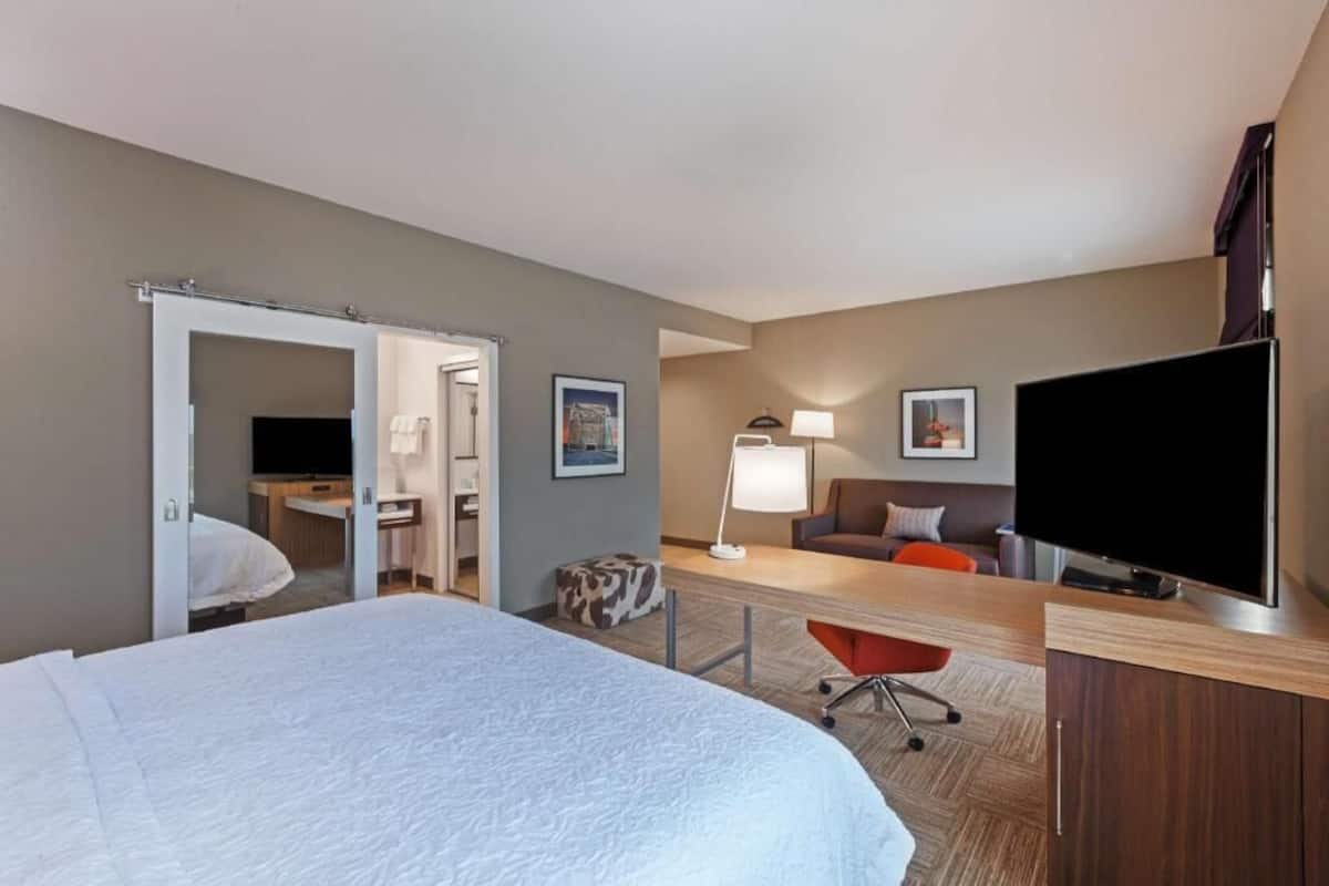 A hotel room with a bed, a desk with a flat-screen TV, a chair, a lamp, and a sofa.