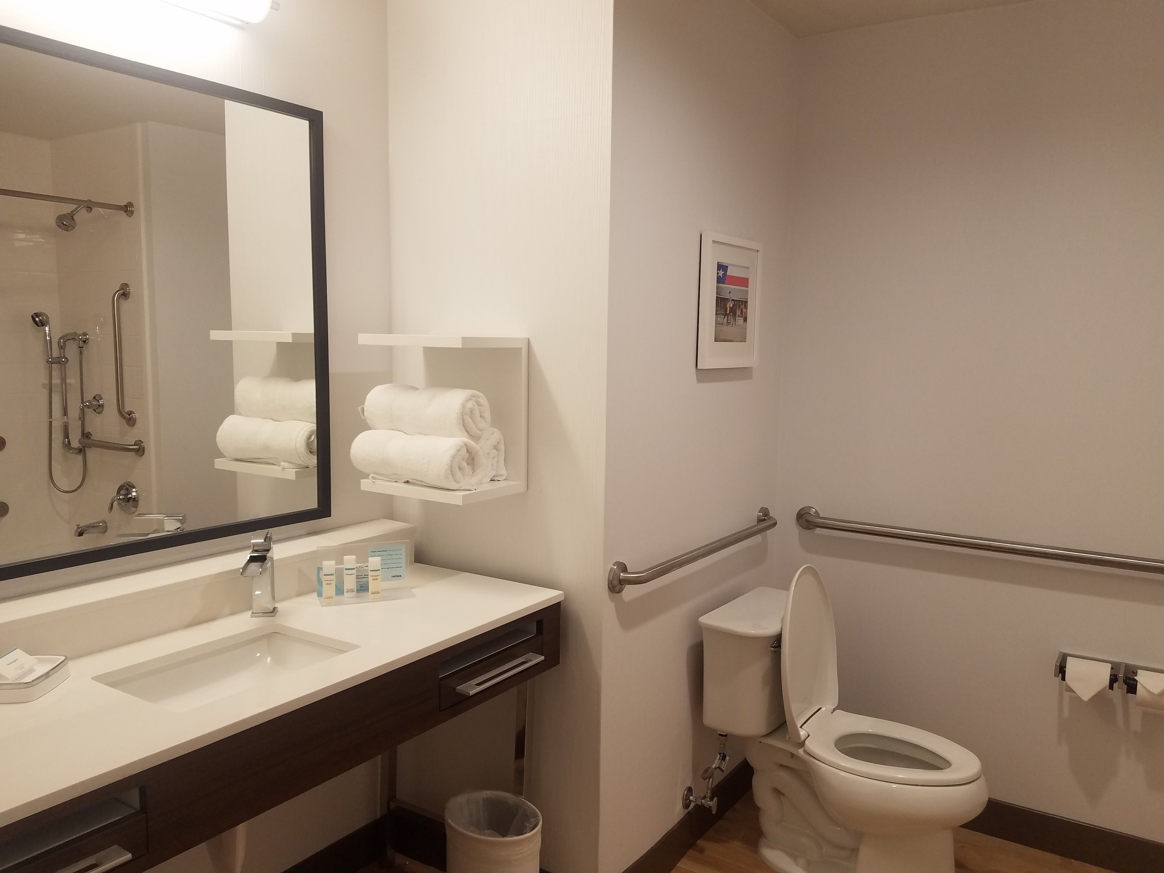 A modern bathroom with a white sink, mirror, and toilet. There are towels on a shelf, a handrail, and a trash can.