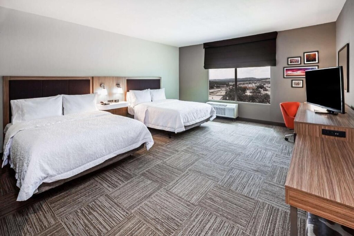 A hotel room with two beds, a desk, a TV, and a window with a city view.