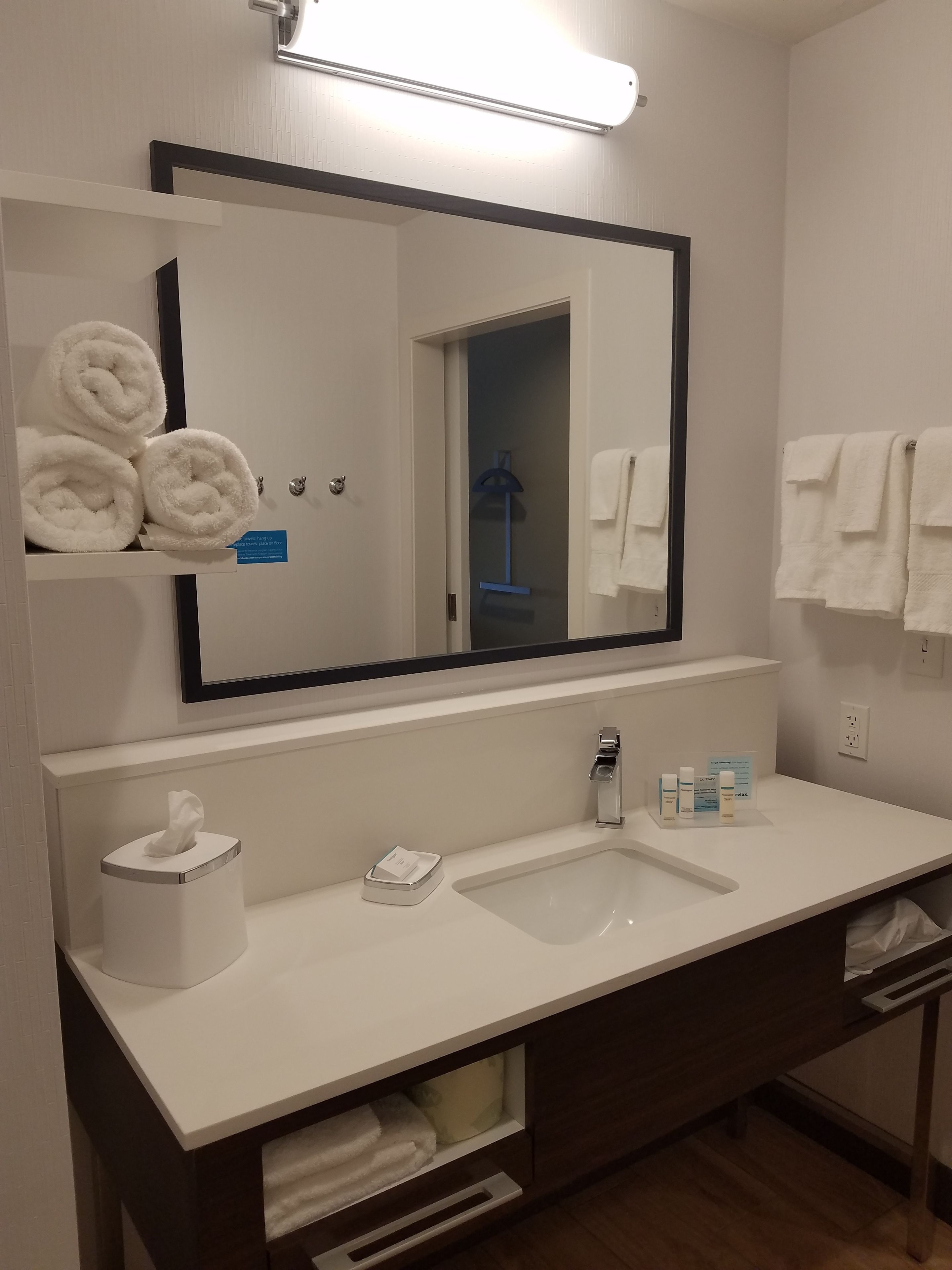 A modern bathroom with a large mirror, white sink, towel rack, and a tissue dispenser.