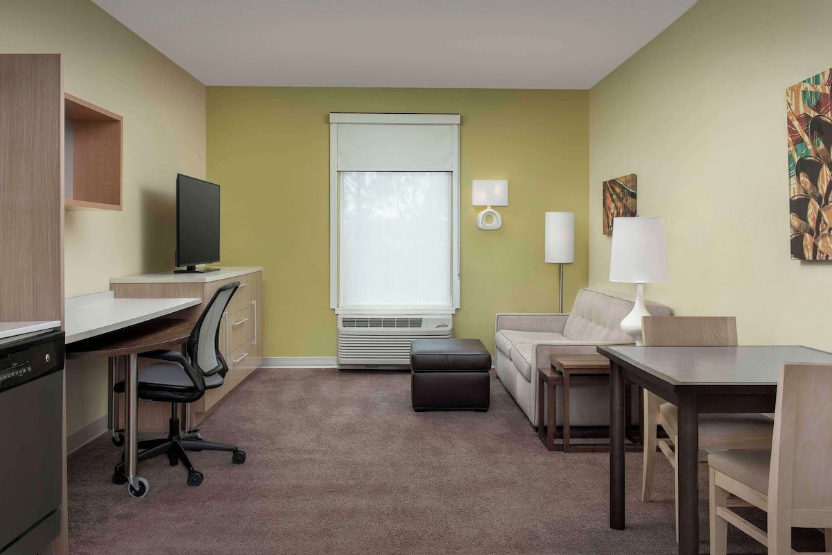 A compact hotel room with a desk, chair, sofa, and a small dining table.