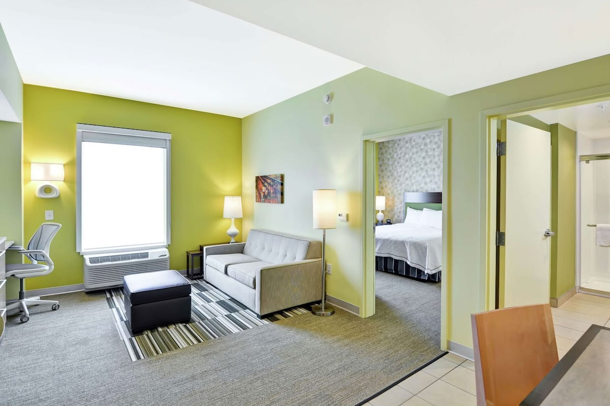 A hotel room with a sofa, armchair, bedside table, lamp, and a view of the bathroom.