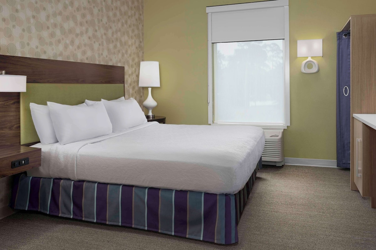 A hotel room with a neatly made bed, a nightstand with a lamp, a window with blinds, and a wooden wardrobe.