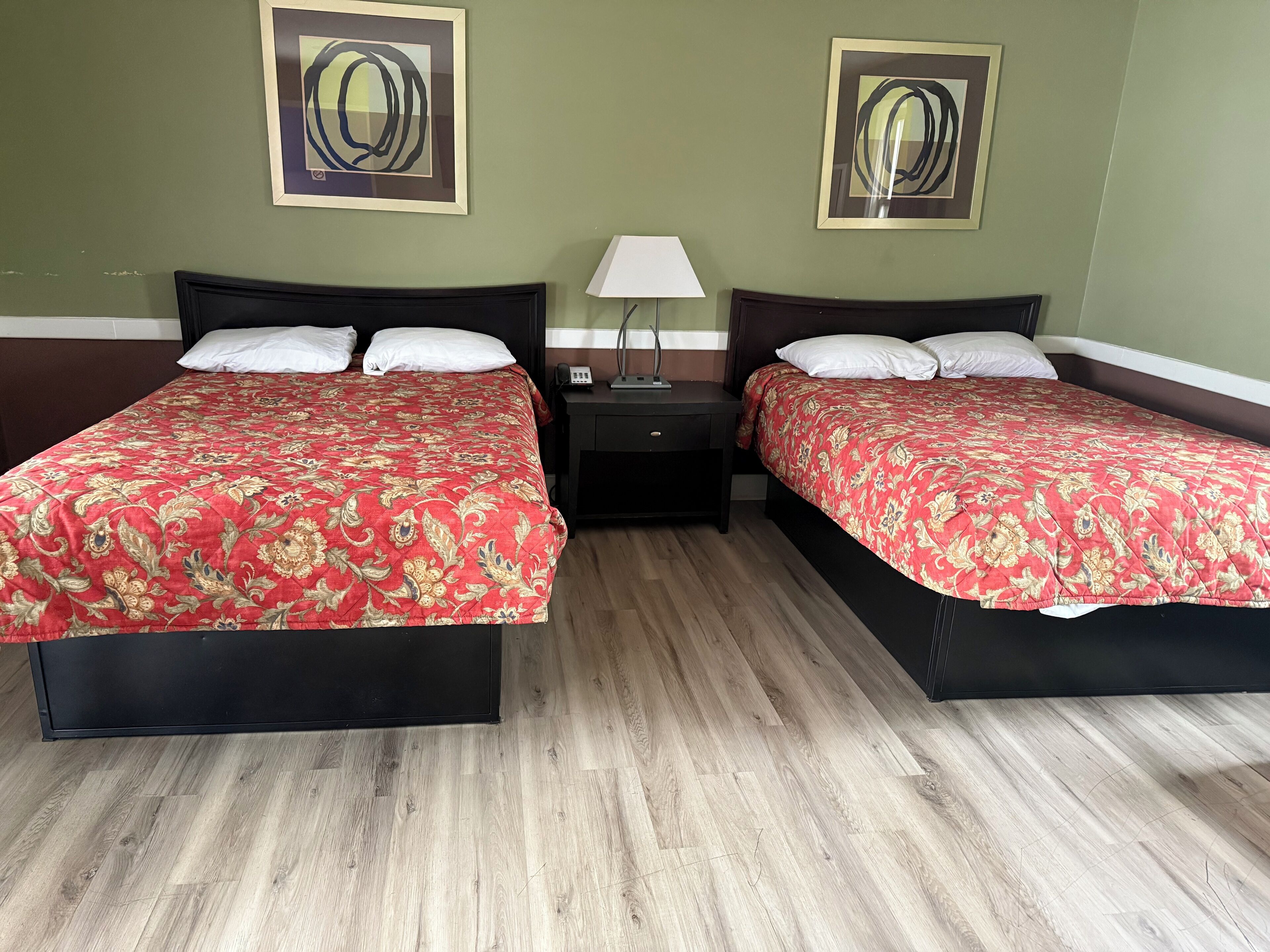 Ein Hotelzimmer mit zwei Betten, jeweils mit einer roten, floralen Tagesdecke, einem Nachttisch mit Lampe und zwei abstrakten Bildern an den Wänden.