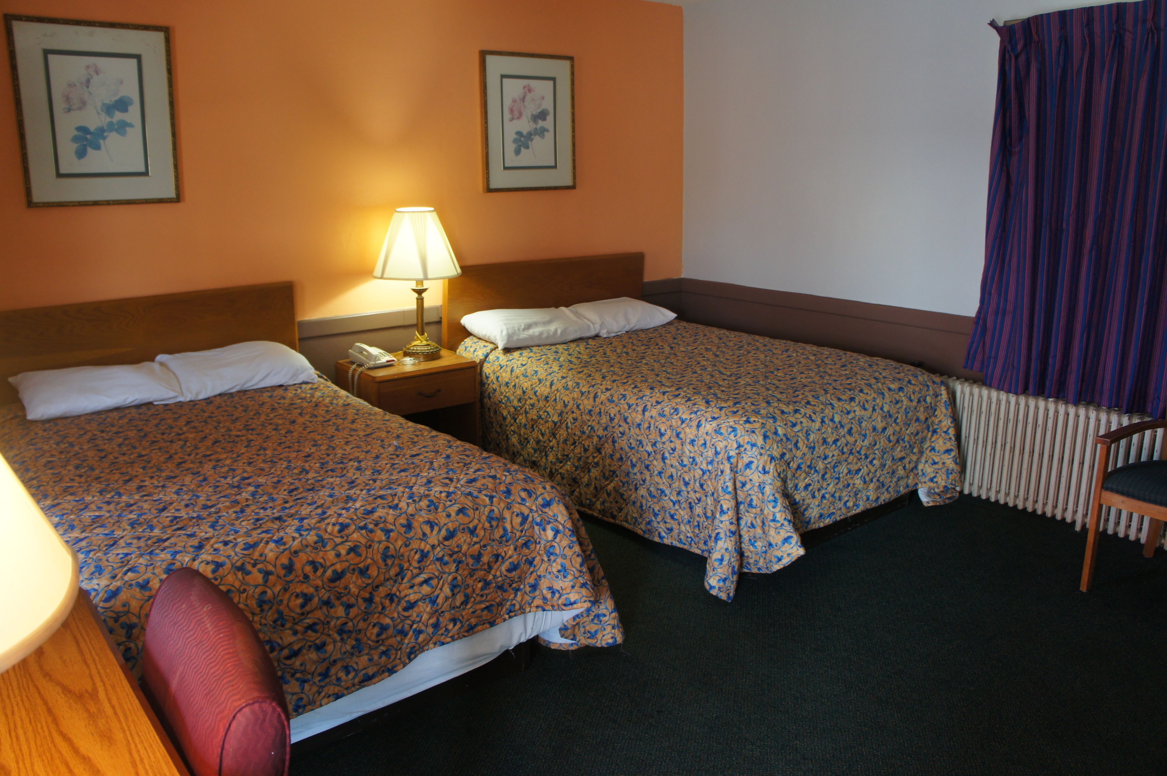 Una camera d'albergo con due letti, un comodino con una lampada, una sedia e un termosifone.