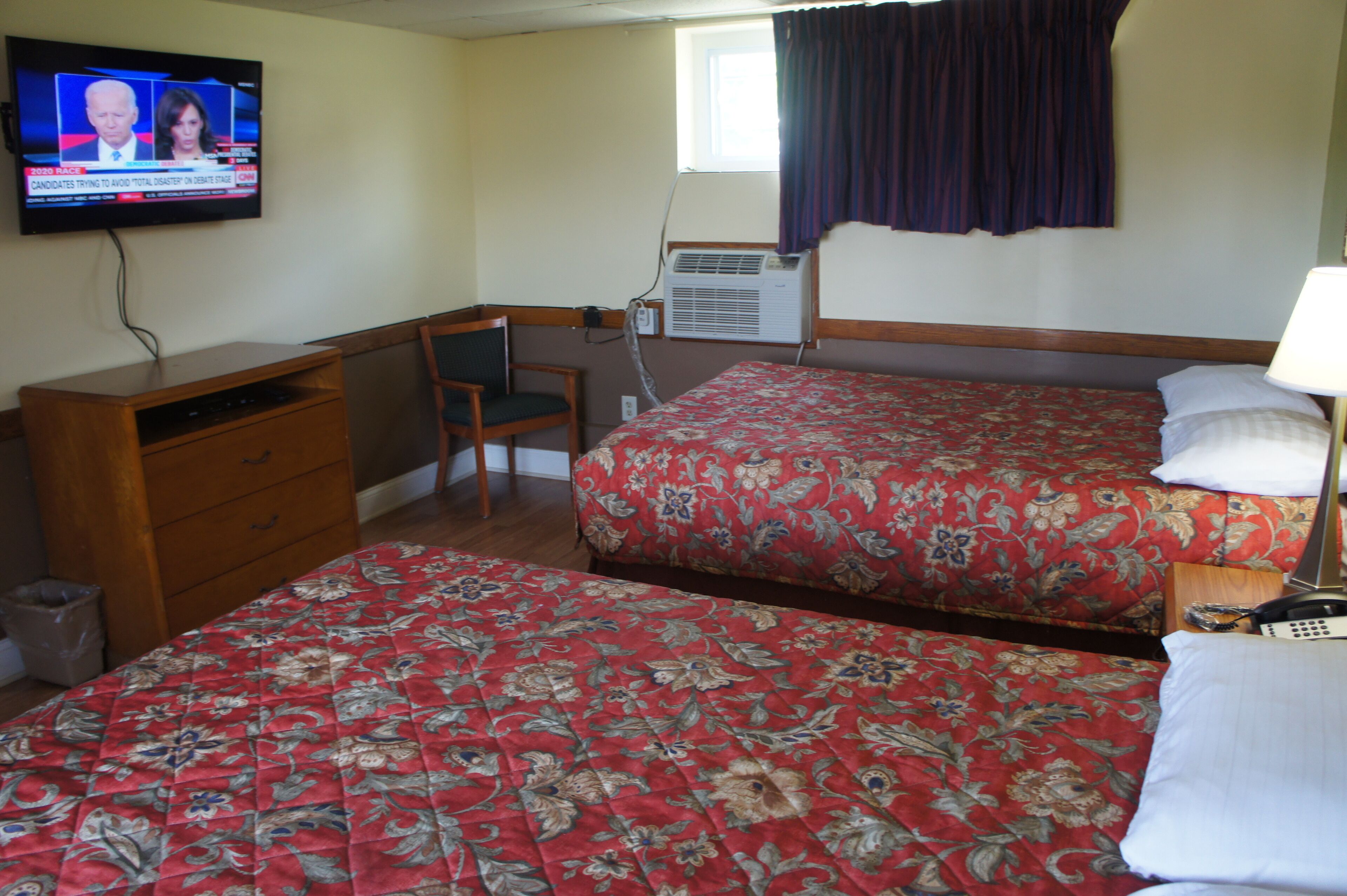 A hotel room with two beds, a desk, a chair, a TV, and an air conditioning unit.