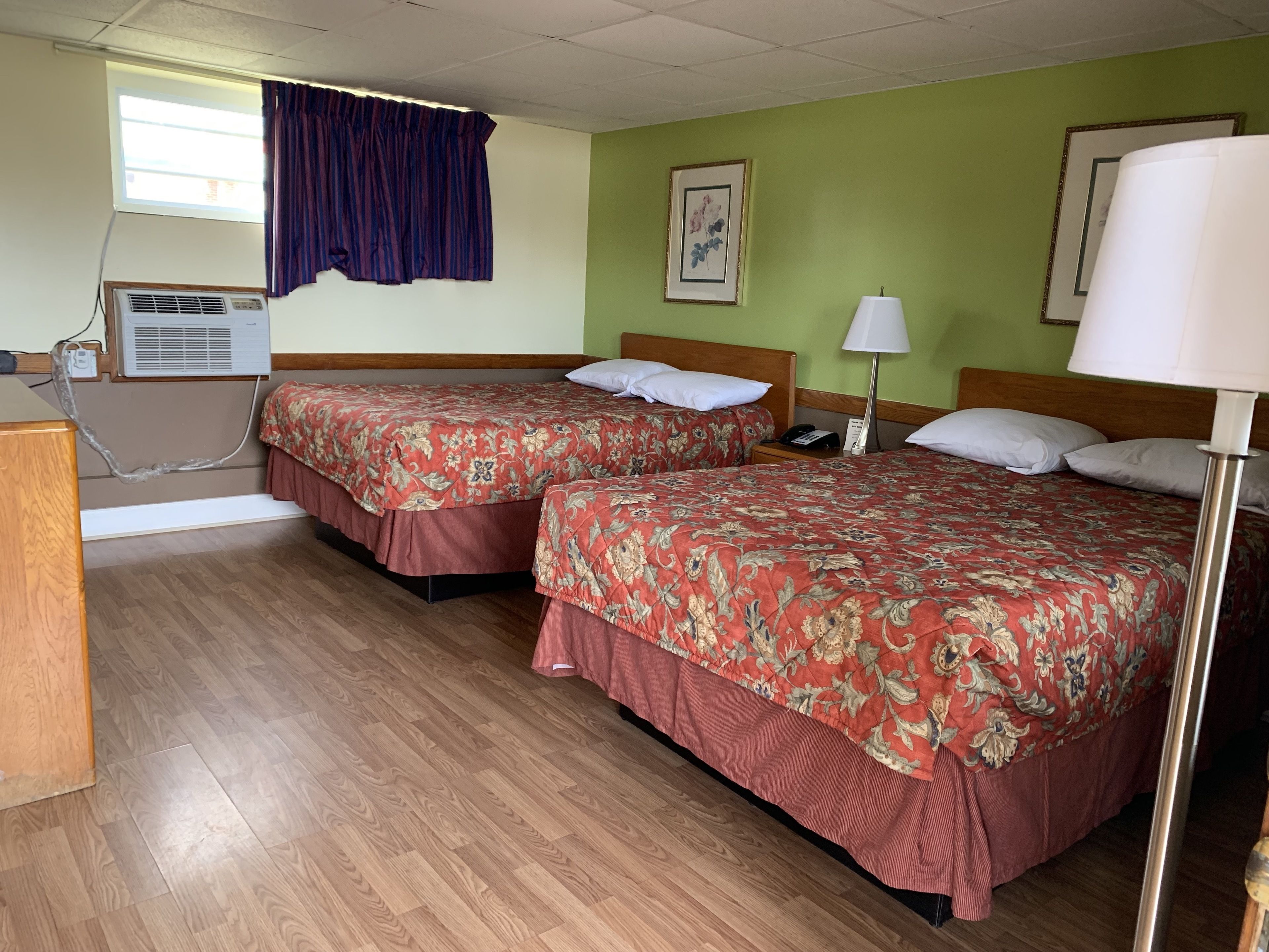 A hotel room with two beds, a desk, an air conditioning unit, a lamp, and a framed picture on the wall.