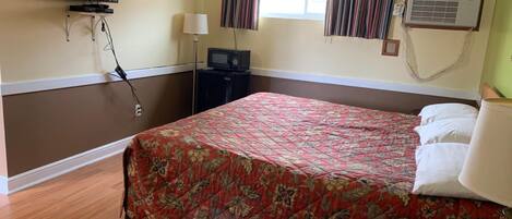 A hotel room with a bed, a television mounted on the wall, a desk with a telephone, and an air conditioning unit.