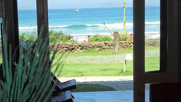 A view from a room with a large window overlooking a beach, a stone wall, and a chair outside. - Casa na Beira da Praia de Geribá (Búzios)