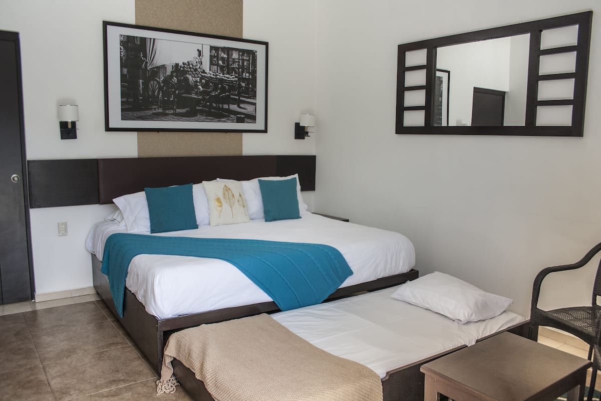 Habitación de hotel ordenada con una cama grande, otra más pequeña, una silla, una mesita de noche, un espejo en la pared y un cuadro enmarcado sobre la cama.