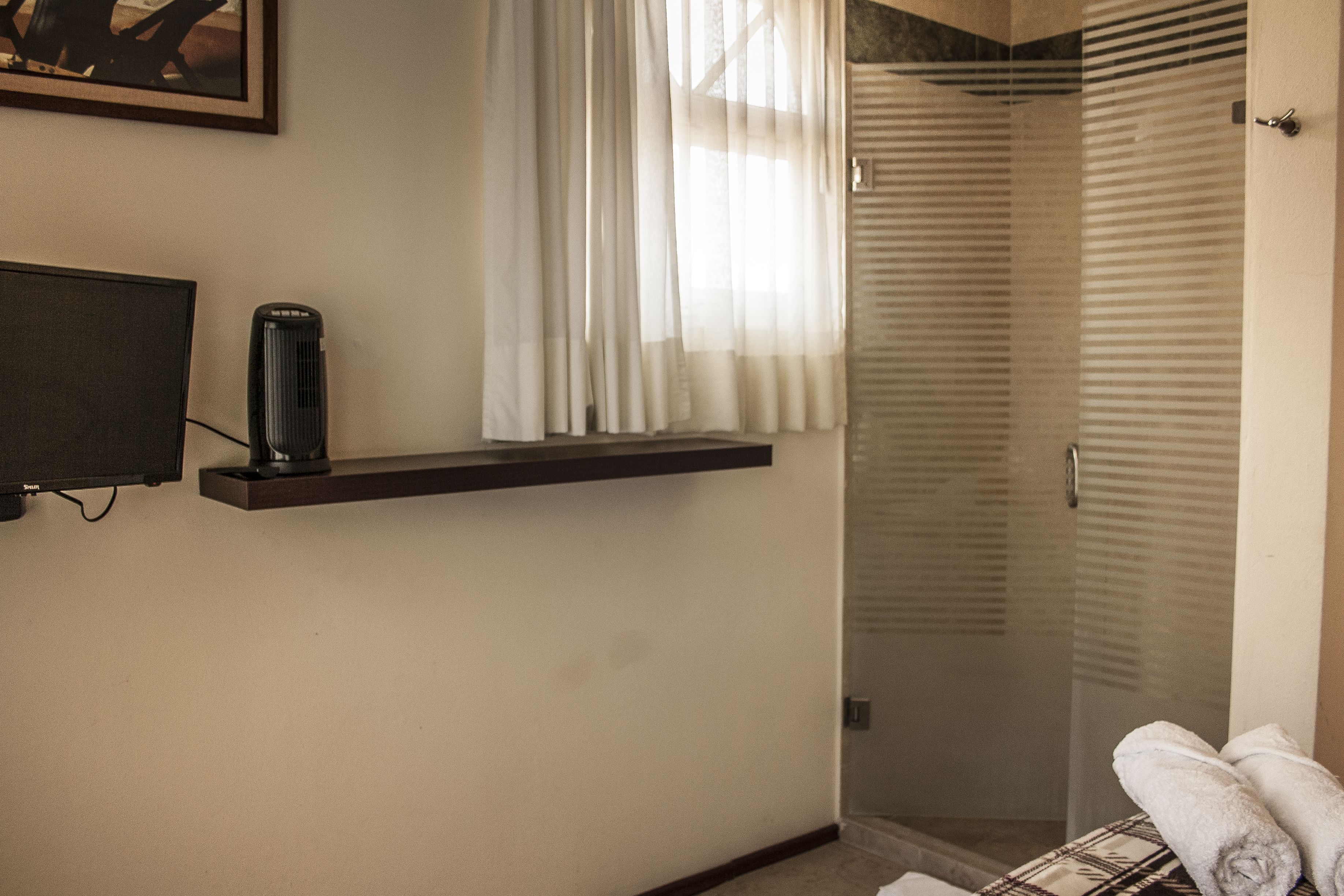 A hotel room with a TV on a shelf, a window with sheer curtains, a shower area, and a towel on the floor.