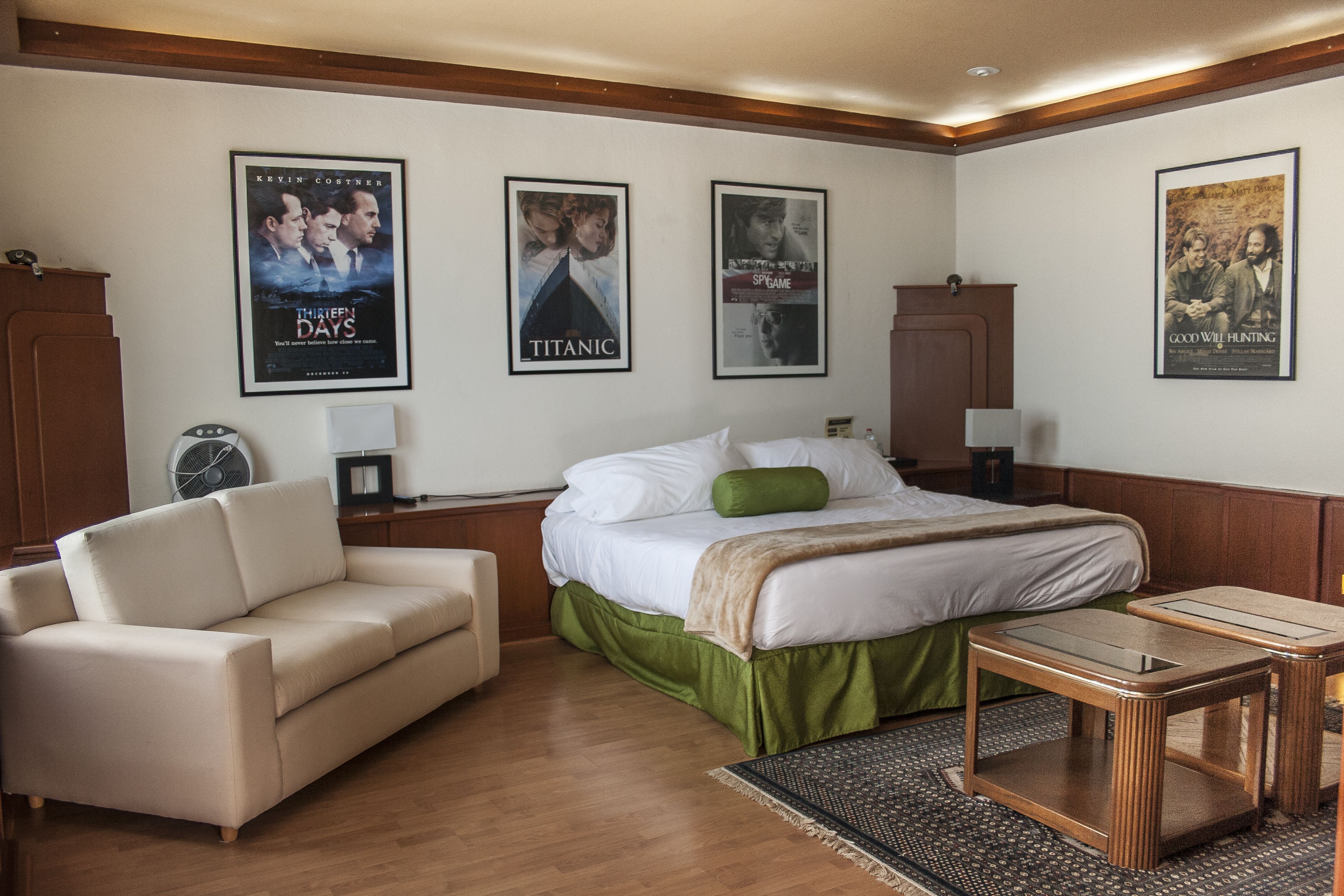 A hotel room with a bed, a green bed skirt, a beige sofa, a wooden nightstand, and a table with a glass top.
