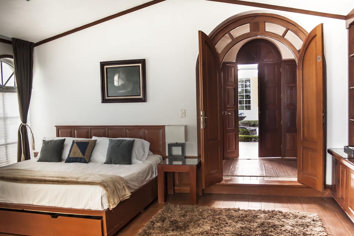Un dormitorio con cama, una mesita de noche, una alfombra, un cuadro enmarcado en la pared y una puerta abierta que da a un balcón.