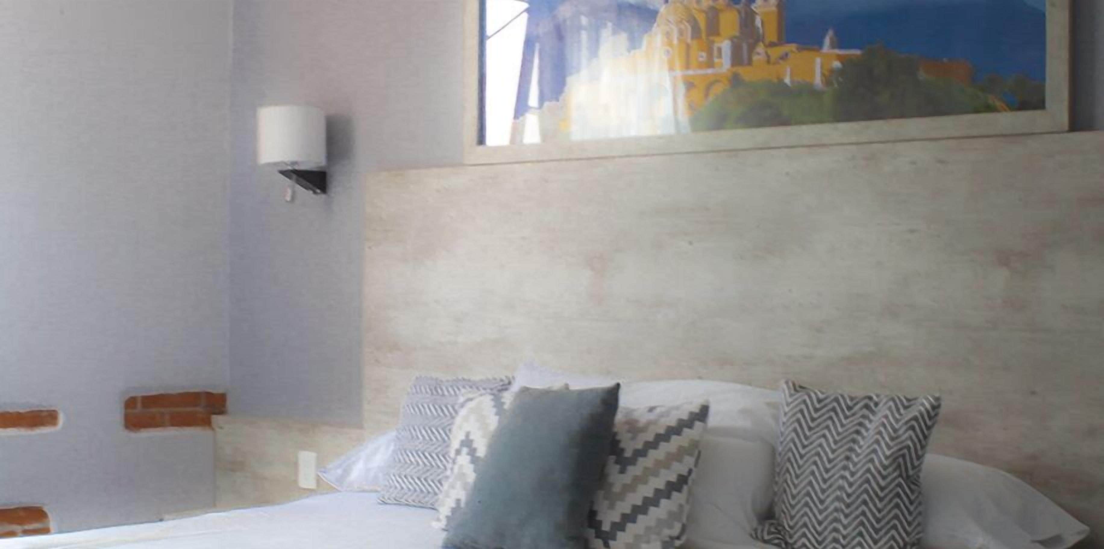 A hotel room with a bed, patterned pillows, a wall-mounted light, and a painting of a building.