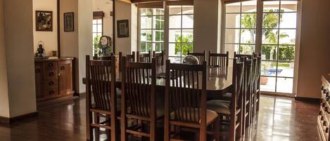 A dining area with a wooden table and chairs, large windows with a view of greenery, and a wooden cabinet with drawers.