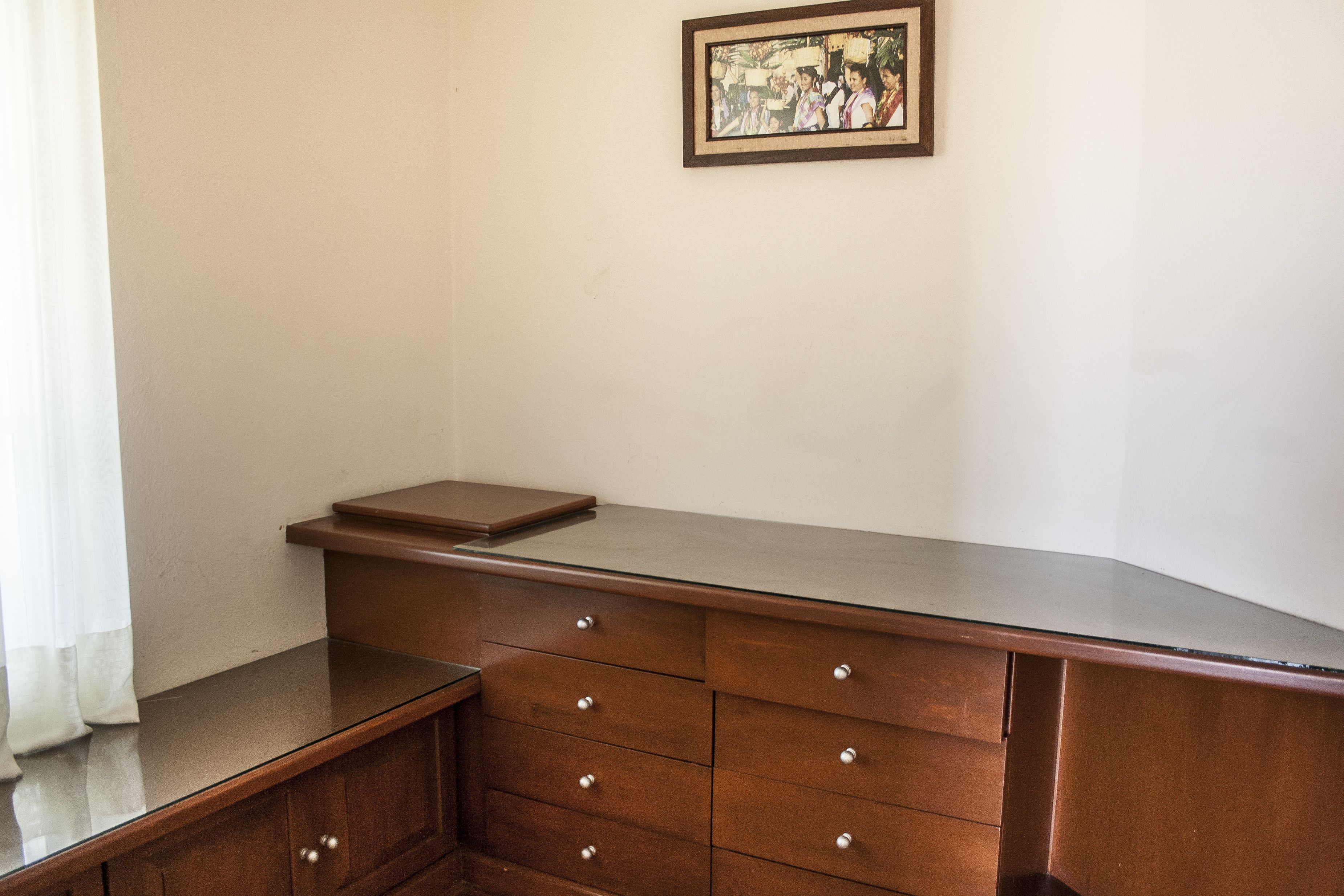 A corner of a room with a wooden desk, a framed picture on the wall, and a window with curtains.