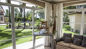 Une pièce dotée d’une grande porte-fenêtre coulissante donnant sur un jardin, d’un canapé avec des coussins, d’une petite table sur laquelle est posée une plante, et d’une étagère en bois où sont rangées des serviettes.