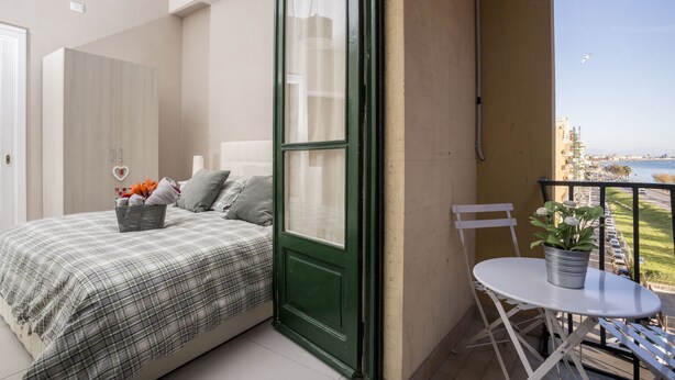A balcony with a view of the sea, a bed with a plaid bedspread, a small table with a potted plant, and a chair. - B&B Monica's House (Salerno)